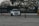 A white car is drifting on a wide, open area, surrounded by large trees in the background. Orange traffic cones are placed sporadically around the scene, suggesting an organized driving or racing course.