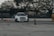 A white car is drifting on a wide, open area, surrounded by large trees in the background. Orange traffic cones are placed sporadically around the scene, suggesting an organized driving or racing course.
