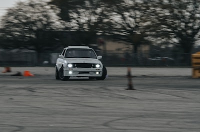 A white car is drifting on a wide, open area, surrounded by large trees in the background. Orange traffic cones are placed sporadically around the scene, suggesting an organized driving or racing course.