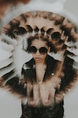 A surreal and artistic portrait features a person wearing sunglasses and multiple layered necklaces. The image creates a kaleidoscope effect with repeated reflections of the person's face, adding a unique, almost dream-like quality. The person wears a black leather jacket and their expression is calm and self-assured.