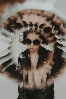 A surreal and artistic portrait features a person wearing sunglasses and multiple layered necklaces. The image creates a kaleidoscope effect with repeated reflections of the person's face, adding a unique, almost dream-like quality. The person wears a black leather jacket and their expression is calm and self-assured.