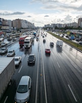 A multi-lane highway with numerous vehicles, including cars and trucks, travels in both directions. The road surface appears wet from recent rain, reflecting the overcast sky. Tall apartment buildings line one side of the highway, while smaller commercial establishments are visible on the other side. Light traffic suggests a period of urban transit.