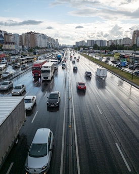 A multi-lane highway with numerous vehicles, including cars and trucks, travels in both directions. The road surface appears wet from recent rain, reflecting the overcast sky. Tall apartment buildings line one side of the highway, while smaller commercial establishments are visible on the other side. Light traffic suggests a period of urban transit.