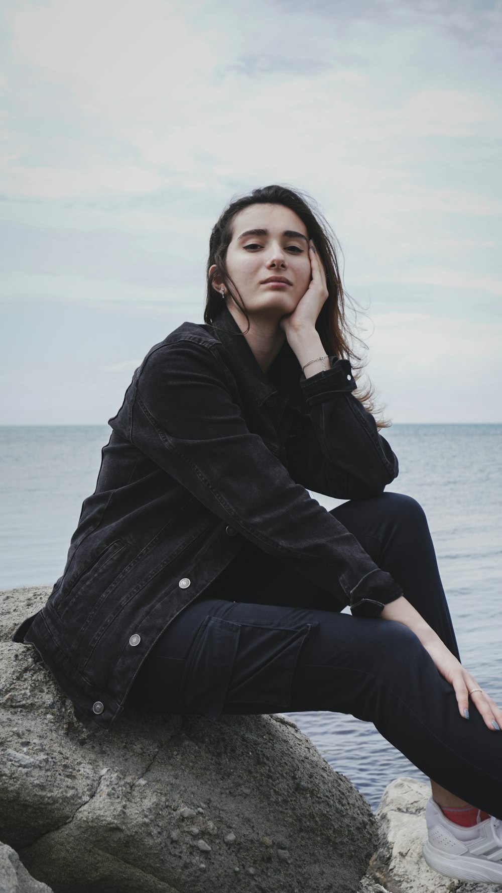 Frau im schwarzen Mantel sitzt tagsüber auf Felsen in der Nähe des Meeres
