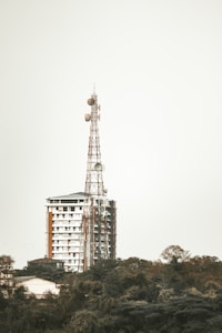 A tall communication tower stands prominently beside a modern multi-story building. The structure is set against a cloudy sky and the building appears newly constructed with a combination of white and brown exterior finishes. In the foreground, lush green foliage adds a natural element to the scene.