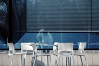 A row of white chairs and tables is set up outside a building with large reflective windows. A person appears to be cleaning or moving an object inside the building, partially visible through the glass. The blinds are drawn down halfway, casting shadows and adding a sense of enclosure.