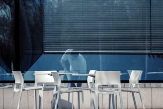 A row of white chairs and tables is set up outside a building with large reflective windows. A person appears to be cleaning or moving an object inside the building, partially visible through the glass. The blinds are drawn down halfway, casting shadows and adding a sense of enclosure.