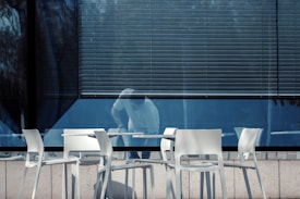A row of white chairs and tables is set up outside a building with large reflective windows. A person appears to be cleaning or moving an object inside the building, partially visible through the glass. The blinds are drawn down halfway, casting shadows and adding a sense of enclosure.
