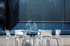 A row of white chairs and tables is set up outside a building with large reflective windows. A person appears to be cleaning or moving an object inside the building, partially visible through the glass. The blinds are drawn down halfway, casting shadows and adding a sense of enclosure.