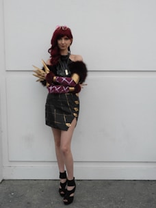 A person in a cosplay outfit poses against a plain background. The outfit features a dark, off-shoulder top with fur accents and a plaid skirt with gold details. The individual wears a red wig and has accessorized with statement pieces like golden spiked armwear, purple gloves, and high-heeled shoes.