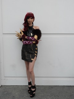 A person in a cosplay outfit poses against a plain background. The outfit features a dark, off-shoulder top with fur accents and a plaid skirt with gold details. The individual wears a red wig and has accessorized with statement pieces like golden spiked armwear, purple gloves, and high-heeled shoes.