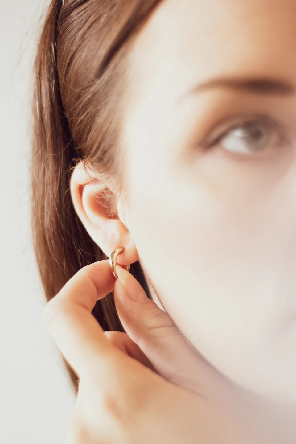 The image shows a close-up view of a person's face, focusing on their ear and the gold hoop earring they are wearing. The earring is positioned at the top of the earlobe, adding a touch of elegance to the scene. The background appears to be a blurred gray color, which contrasts with the golden hue of the earring and accentuates its presence in the image.