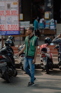A man is standing on the street wearing a green vest and blue jeans. He appears to be managing or directing motorbike parking. Several parked motorbikes are seen around him. In the background, there are people and a large sign displaying prices in an outdoor market or shop area. Various items, including backpacks, are visible on display.