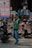 A man is standing on the street wearing a green vest and blue jeans. He appears to be managing or directing motorbike parking. Several parked motorbikes are seen around him. In the background, there are people and a large sign displaying prices in an outdoor market or shop area. Various items, including backpacks, are visible on display.