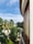 A curved balcony with a glass railing overlooks a lush garden filled with various palm trees and greenery. The sky above is partly cloudy, showing patches of blue. On the right, there's a building with wooden window shutters, adding a tropical feel to the setting.