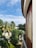 A curved balcony with a glass railing overlooks a lush garden filled with various palm trees and greenery. The sky above is partly cloudy, showing patches of blue. On the right, there's a building with wooden window shutters, adding a tropical feel to the setting.