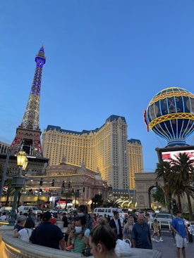 A bustling city scene with a replica of the Eiffel Tower, a brightly lit hot air balloon sign, and a grand hotel building set against a twilight sky. Numerous people are walking around, some wearing masks, and there is a vibrant mix of lights and activity.