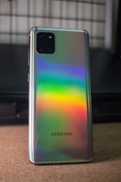 A smartphone with a reflective back surface displays a spectrum of rainbow colors. The brand name is visible near the bottom, and there is a dual-camera module at the top left corner. The background is blurred, focusing attention on the phone.