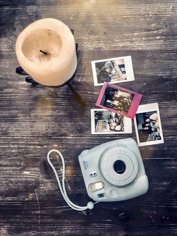 A vintage-style camera rests on a rustic wooden surface next to three instant photos and a large, unlit pillar candle. The photos appear to show people and settings from events or gatherings, and there is a gentle light effect across the scene.