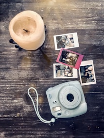 A vintage-style camera rests on a rustic wooden surface next to three instant photos and a large, unlit pillar candle. The photos appear to show people and settings from events or gatherings, and there is a gentle light effect across the scene.