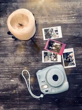 A vintage-style camera rests on a rustic wooden surface next to three instant photos and a large, unlit pillar candle. The photos appear to show people and settings from events or gatherings, and there is a gentle light effect across the scene.