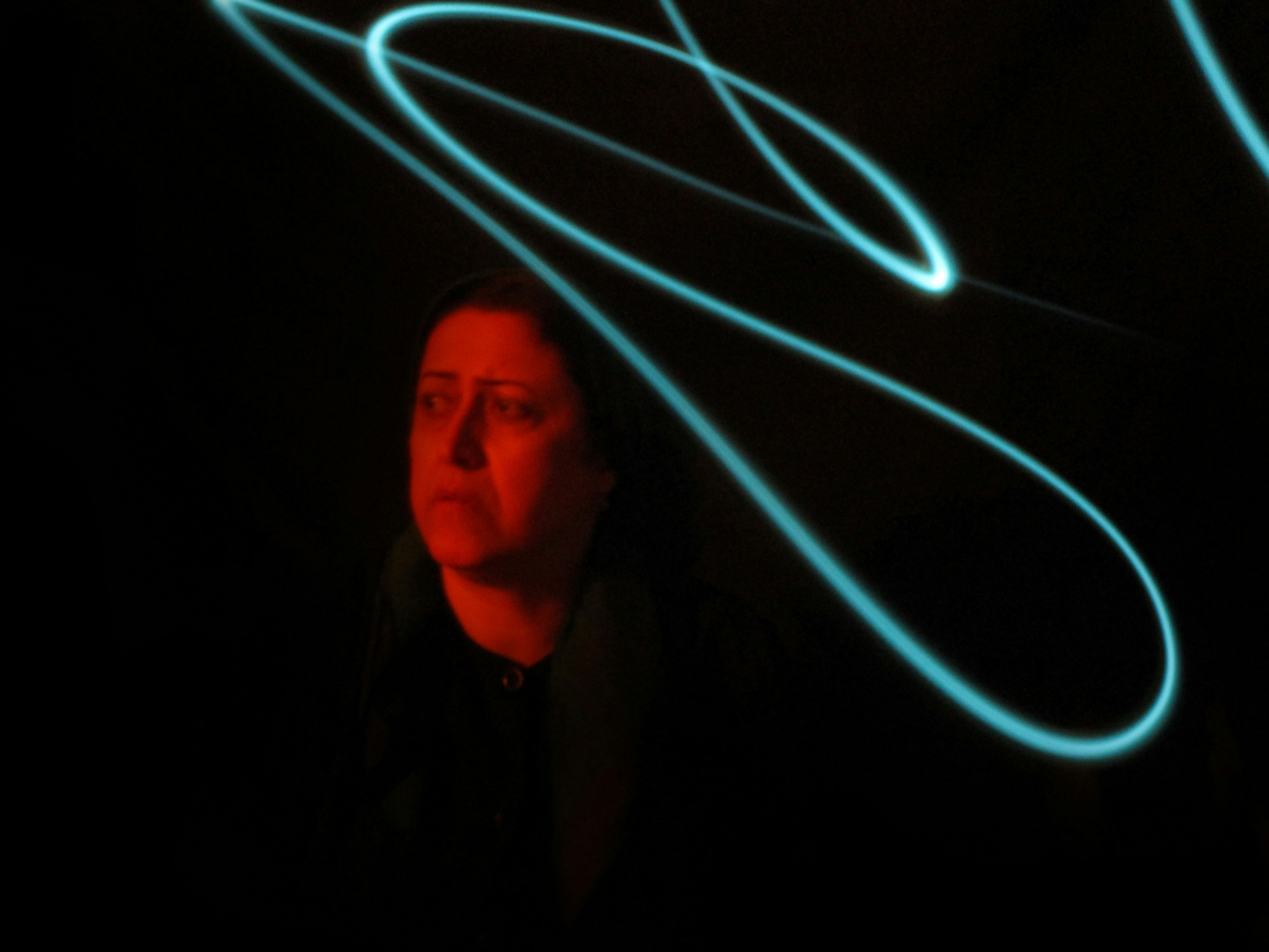 A lone, somber figure stands in near-darkness as turquoise light trails swirl above, creating a surreal, abstract ambience.