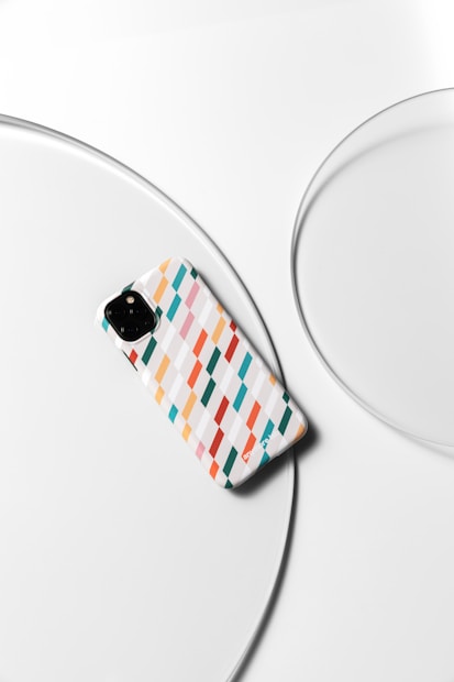 A smartphone with a colorful, geometric-patterned case featuring diagonal stripes in shades of red, orange, blue, and white, placed on a sleek, round white surface with concentric circular designs.