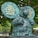 A large greenish-blue statue of a cartoon-like anthropomorphic animal wearing a necktie and belt, standing with chest puffed out. It features two speech bubbles with French text. The statue is set against a backdrop of green trees.