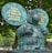 A large greenish-blue statue of a cartoon-like anthropomorphic animal wearing a necktie and belt, standing with chest puffed out. It features two speech bubbles with French text. The statue is set against a backdrop of green trees.