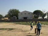 Several people are scattered across a dusty field, apparently engaged in a game or casual activity. One person in a blue shirt and yellow cap stands near a stack of bricks arranged in a column. Another person in a green shirt appears to be involved in the activity near the stack of bricks. The background includes an old, single-story building with a central entrance and rectangular windows. There are some trees with green foliage around the perimeter of the field, and a clear blue sky overhead.