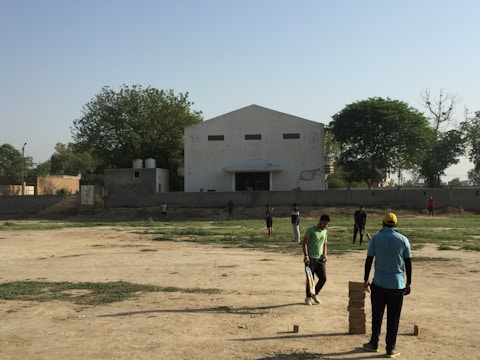 Several people are scattered across a dusty field, apparently engaged in a game or casual activity. One person in a blue shirt and yellow cap stands near a stack of bricks arranged in a column. Another person in a green shirt appears to be involved in the activity near the stack of bricks. The background includes an old, single-story building with a central entrance and rectangular windows. There are some trees with green foliage around the perimeter of the field, and a clear blue sky overhead.
