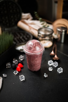 A berry smoothie in a clear plastic cup with a domed lid is placed on a dark countertop. Surrounding the smoothie are scattered ice cubes and fresh raspberries and blackberries. In the background, there are kitchen items including a waffle iron, a glass jar, and metal utensils.