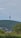 A tall telecommunications tower rises from a densely forested hill, surrounded by lush greenery. A small town with various buildings is visible at the base of the hill, and overhead, a clear blue sky occupies the background.