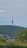 A tall telecommunications tower rises from a densely forested hill, surrounded by lush greenery. A small town with various buildings is visible at the base of the hill, and overhead, a clear blue sky occupies the background.