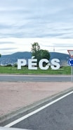 Large white letters spelling 'PÉCS' are prominently displayed on a grassy area with a backdrop of green hills and a television tower. A partly cloudy sky is visible in the background, and there are trees surrounding the letters. A road with a traffic sign is present in the foreground.