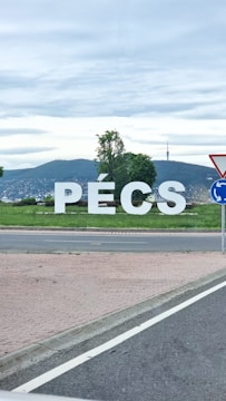 Large white letters spelling 'PÉCS' are prominently displayed on a grassy area with a backdrop of green hills and a television tower. A partly cloudy sky is visible in the background, and there are trees surrounding the letters. A road with a traffic sign is present in the foreground.