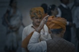 A group of people with one visible from the back wearing an orange turban, raising their hand. Another person in the background also wearing a similar patterned turban appears slightly out of focus.