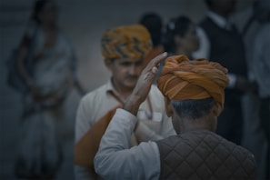 A group of people with one visible from the back wearing an orange turban, raising their hand. Another person in the background also wearing a similar patterned turban appears slightly out of focus.