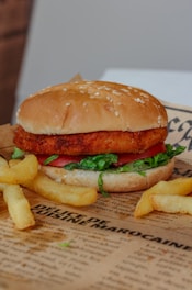 A crispy chicken burger sits on a vintage-style paper, adorned with sesame seeds on a fluffy bun. Inside, layers of fresh tomato and lettuce are visible, creating a delicious contrast with the golden-brown chicken fillet. Crispy french fries accompany the burger, enhancing the appetizing presentation.