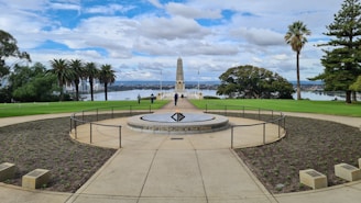 A memorial site with a central obelisk set against a backdrop of trees and a body of water. The area is landscaped with grass and plants, accompanied by palm trees and other greenery. The sky is filled with clouds, and there are a few people walking around the site.