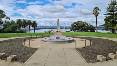 A memorial site with a central obelisk set against a backdrop of trees and a body of water. The area is landscaped with grass and plants, accompanied by palm trees and other greenery. The sky is filled with clouds, and there are a few people walking around the site.