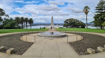 A memorial site with a central obelisk set against a backdrop of trees and a body of water. The area is landscaped with grass and plants, accompanied by palm trees and other greenery. The sky is filled with clouds, and there are a few people walking around the site.