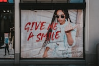 A fashion advertisement features a person with long braided hair wearing sunglasses and a denim outfit. The individual is making a bold gesture with their hand while standing against a wooden wall. The text 'GIVE ME A SMILE' is prominently displayed in red, along with a hashtag and the website URL 'tally-weijl.com'.