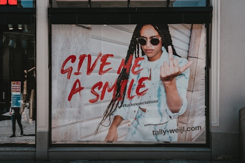 A fashion advertisement features a person with long braided hair wearing sunglasses and a denim outfit. The individual is making a bold gesture with their hand while standing against a wooden wall. The text 'GIVE ME A SMILE' is prominently displayed in red, along with a hashtag and the website URL 'tally-weijl.com'.