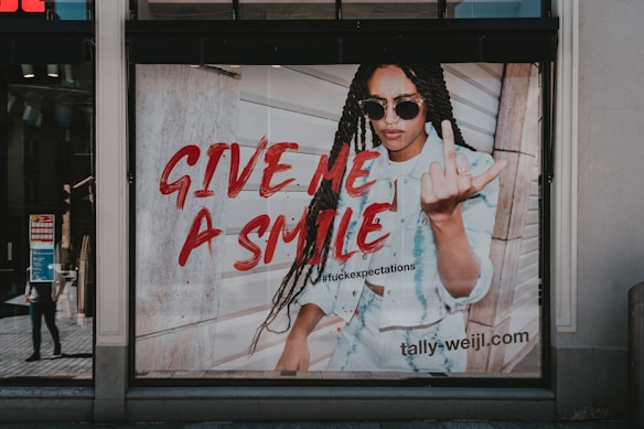 A fashion advertisement features a person with long braided hair wearing sunglasses and a denim outfit. The individual is making a bold gesture with their hand while standing against a wooden wall. The text 'GIVE ME A SMILE' is prominently displayed in red, along with a hashtag and the website URL 'tally-weijl.com'.