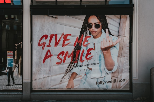 A fashion advertisement features a person with long braided hair wearing sunglasses and a denim outfit. The individual is making a bold gesture with their hand while standing against a wooden wall. The text 'GIVE ME A SMILE' is prominently displayed in red, along with a hashtag and the website URL 'tally-weijl.com'.
