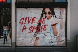 A fashion advertisement features a person with long braided hair wearing sunglasses and a denim outfit. The individual is making a bold gesture with their hand while standing against a wooden wall. The text 'GIVE ME A SMILE' is prominently displayed in red, along with a hashtag and the website URL 'tally-weijl.com'.