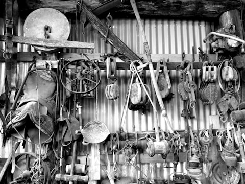 A collection of old tools and equipment hanging on a corrugated metal wall. Includes pulleys, a saddle, various hand tools, and industrial items made of wood and metal.