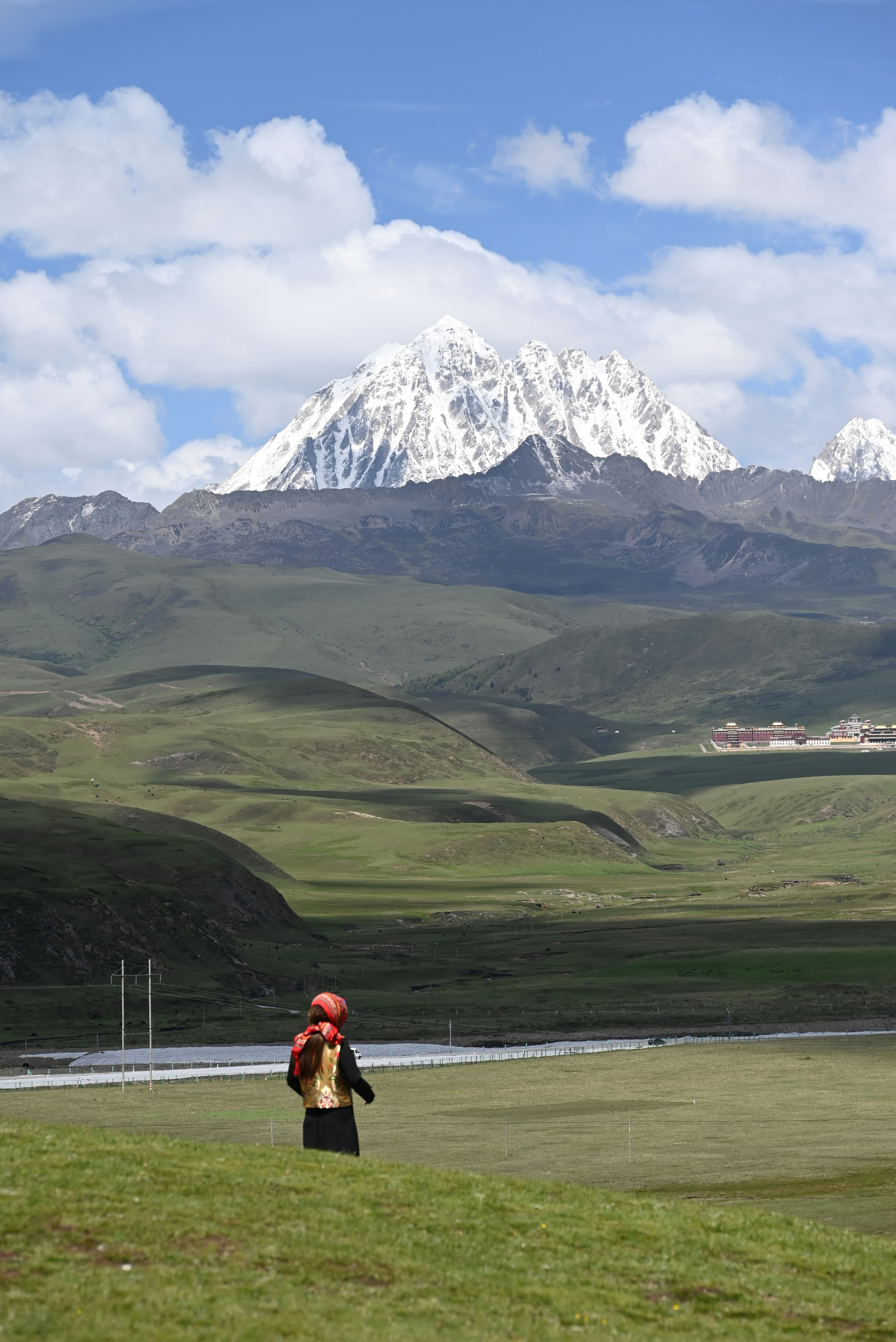 A vast, green landscape stretches out under a bright blue sky dotted with fluffy clouds. A snow-capped mountain dominates the background, while in the foreground, a person wearing traditional attire with a red headscarf stands gazing towards the mountains.