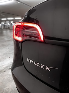 A close-up of the rear part of a dark-colored car with an illuminated red tail light. The vehicle features a 'SpaceX' logo prominently displayed on its back. It is parked in an indoor garage with other parking spaces visible in the background.